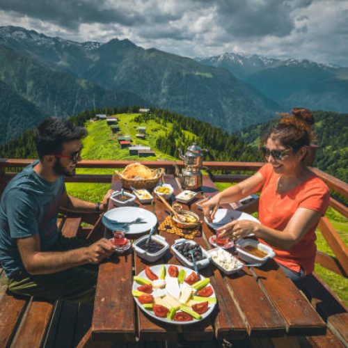 A young tourist couple eating delicious Turkish breakfast with various colorful foods and Black Sea Region food stretch muhlama or kuymak (Turkish Cheese Fondue) with nature landscape view at Pokut Highland in Çamlıhemşin, Rize in Karadeniz region of Türkiye