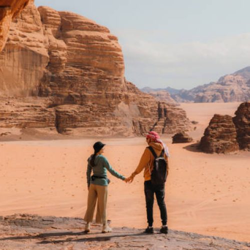Young heterosexual married couple staying at the edge of the cliff looking at the red sands and the mountains of Jordan